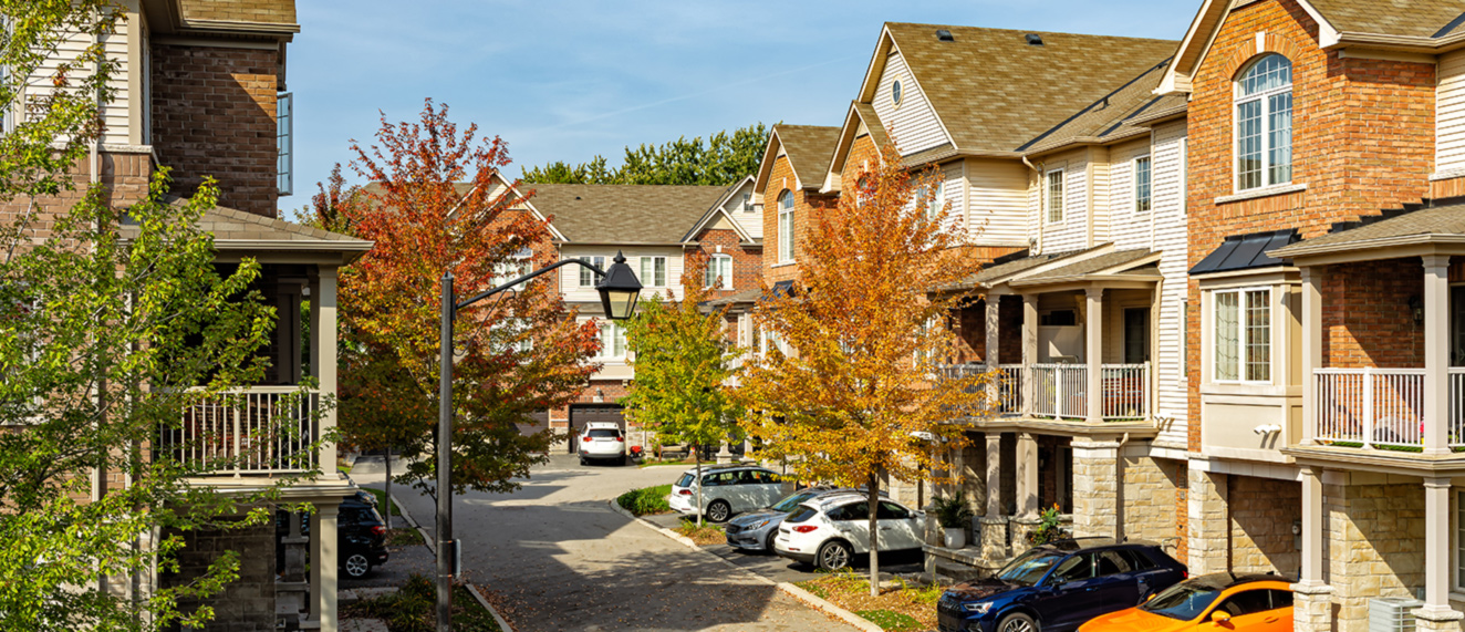 The image features a row of houses lined along a street in a residential area. It showcases elements typical of a neighborhood, including parked cars and autumn trees, creating a picturesque urban setting. The scene is enhanced by clear skies, emphasizing the homes and surrounding environment.