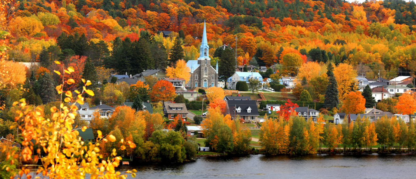 Colorful autumn foliage surrounds a small lakeside village with a prominent church and a backdrop of dense forest.