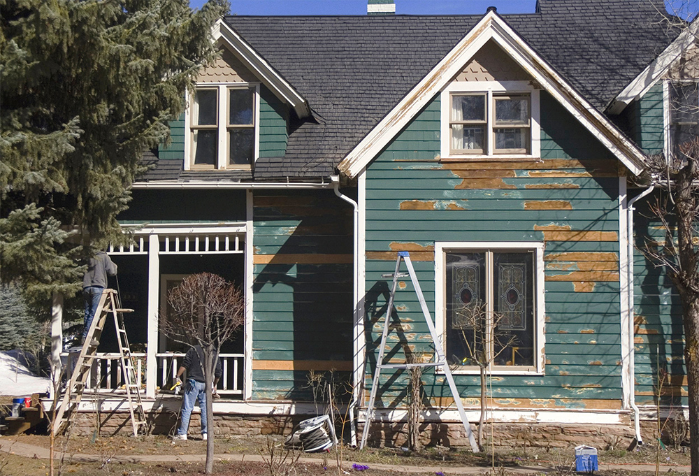 Two people on ladders working on the exterior of an old house with peeling green paint, surrounded by trees.