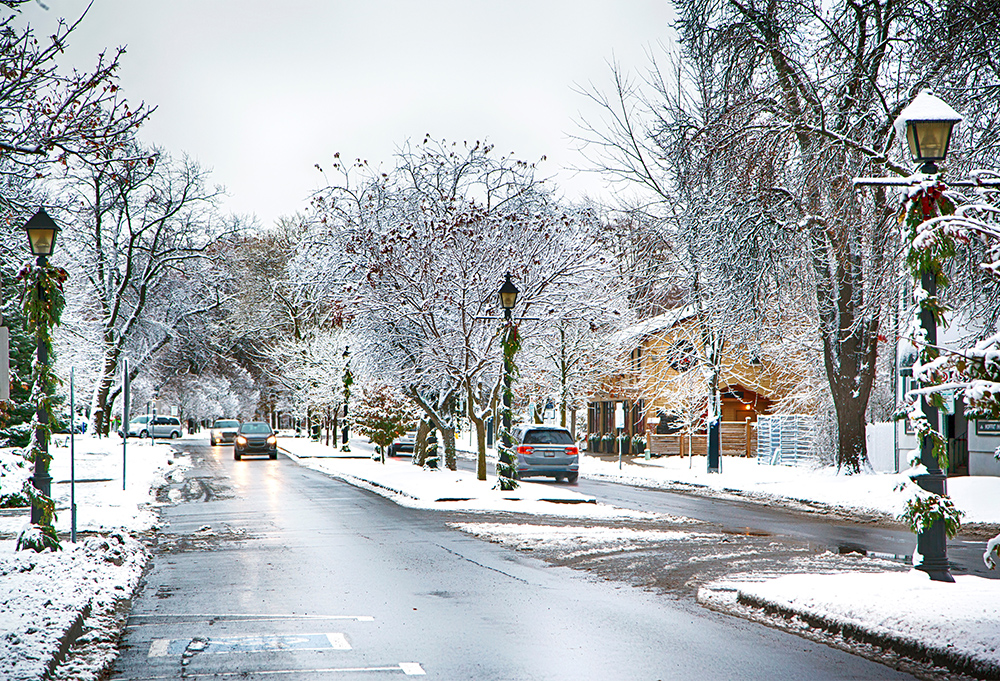 A snowy street scene in winter with snow-covered trees lining the road. Cars are parked and driving along the wet pavement. Street lamps are decorated with green garlands, and houses are visible in the background. The sky is overcast.